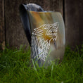 Load image into Gallery viewer, Et poleret Vikingekrus ulvehorn (600-800 ml), med et stiliseret ulvehoveddesign, der er indviklet udskåret på siden, står oprejst på grønt græs. Baggrunden er lavet af træplanker. Dette unikt designede horn viser en blanding af mørke og lyse nuancer.
