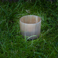 Load image into Gallery viewer, En opretstående Lysestage til fyrfadslys, cylindrisk og gennemskinnelig, placeret på en plet af grønt græs. Lignet til en delikat messingholder virker den tom og har en glat overflade med en let glans.