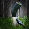 Load image into Gallery viewer, Et mørkt poleret Drikkehorn Ordineret med en gylden rand er støttet oprejst på en lettere, dekorativ hornholder. Opstillingen, der minder om vikingemarkeder, er placeret på grønt græs mod en rustik træbaggrund.