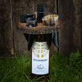 Load image into Gallery viewer, En flaske Hyldemjød mjød, beliggende delvist i græs, er omgivet af et rustikt Holder til vin med horn med sorte buede horn accenter. Det cirkulære træstativ indeholder små horn til servering af drikkevarer og hviler mod en træoverflade i baggrunden.