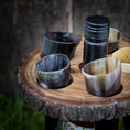 Load image into Gallery viewer, En rustik "Holder til vin med horn" med fem små hornkopper og en sort flaske står på en sløret, jordagtig baggrund. Bakkens naturlige tekstur og jordfarver fremhæver æstetikken, hvilket gør den perfekt til servere drikkevarer.