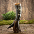 Load image into Gallery viewer, Drikkehorn Yggdrasil U. Mønster, lavet af et poleret dyrehorn, fremviser en tydelig krumning og kommer med et udsmykket udskåret stativ, der ligner det mytiske Yggdrasil-træ. Den rustikke charme ved dette stykke forstærkes yderligere af baggrunden af træplanker og grønt løv, der vækker minder om skatte fundet på et vikingemarked.