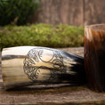 Load image into Gallery viewer, Et udskåret Drikkehorn Yggdrasil U. Mønster hviler på en træoverflade, prydet med indviklede vin- og bladgraveringer, der symboliserer Yggdrasil. Ved siden af står endnu et horn af lignende design. Baggrunden viser en rustik trætekstur med sløret grønt, der fremkalder atmosfæren af et vikingemarked.