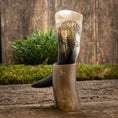 Load image into Gallery viewer, Drikkehorn Yggdrasil U. Mønster og dens stand, begge fremstillet af poleret hornmateriale, er fremvist på en rustik træoverflade med grønt mos og omkringliggende vegetation. Hornet viser en indviklet indgravering af Yggdrasil, der fremkalder den gamle magi, der minder om et vikingemarked.