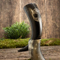 Load image into Gallery viewer, Drikkehorn Yggdrasil U. Mønster er udstillet opretstående på en træstub, omgivet af grønt mos og planter. Dette elegante drikkehorn har indviklede udskæringer med bladmotiver ætset ind i overfladen, der minder om gamle vikingermærkede skatte og påkalder Yggdrasils ånd.