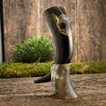 Load image into Gallery viewer, To udsøgte Drikkehorn Yggdrasil U. Mønster, med den ene i et graveret sort design og den anden i en naturlig hornnuance, fremvises stående på en struktureret træoverflade prydet med grønt mos og planter som baggrund.