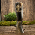 Load image into Gallery viewer, Et poleret Drikkehorn Yggdrasil U. Mønster er lodret placeret på en rustik træoverflade, med grønt mos og græs i baggrunden mod en struktureret trævæg. Scenen udstråler en naturlig og jordnær stemning, der minder om skatte fundet på et vikingemarked.
