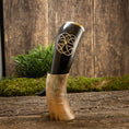 Load image into Gallery viewer, Drikkehorn Templar, et dekorativt vikinge-drikkehorn lavet af polerede bøfler med indviklede udskæringer, hviler oprejst på et træstativ. Baggrunden har en rustik træoverflade prydet med grønt mos og løv, der forstærker den naturlige og historiske æstetik, der minder om vikingemarkeder.