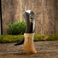 Load image into Gallery viewer, En poleret Drikkehorn Templar står oprejst i en beige meleret holder på en rustik træoverflade. Frisk grønt løv og mos fremhæver de jordiske omgivelser, og hornet har et indviklet udskåret design nær toppen, der minder om dem, man finder på vikingemarkeder.