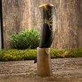 Load image into Gallery viewer, En poleret Drikkehorn Templar med gyldne graveringer hviler i et indviklet formet hornstativ. Opsætningen er placeret på en rustik træflade med en plet grønt og en trævæg i baggrunden, hvilket skaber en middelalderlig æstetik, der minder om vikingemarkeder.