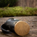 Load image into Gallery viewer, Et nærbillede af Mini Vikingekrus Horn Thors Hammer (100 ml) hviler på siden på en rustik træoverflade. Hornet, der minder om dem, der vises i Roskilde showroom, er lavet af poleret sort materiale med en lys træbase og har en let sløret naturlig grøn baggrund i baggrunden.