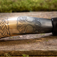Load image into Gallery viewer, Nærbillede af Drikkehorn Yggdrasil med Mønster, der viser dens polerede overflade prydet med indviklede udskæringer. Designet har et sammenflettet knudemønster til venstre, der går over til et træmotiv med detaljerede blade i midten og til højre. Hornet hviler på en rustik træholder, der minder om vikingemarkeder.