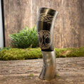 Load image into Gallery viewer, Et dekorativt Drikkehorn Yggdrasil med Mønster, prydet med udskårne designs, står på en træoverflade. Den øverste halvdel viser indviklede keltiske knudemønstre og et stiliseret ugleansigt. Baggrunden, der fremkalder stemningen af vikingemarkeder, omfatter grønt mos og planter, der forstærker dens rustikke, naturlige charme.