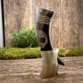 Load image into Gallery viewer, Drikkehorn Yggdrasil med Mønster, prydet med indviklede hvide udskæringer med et knudedesign og et skjold, hviler på en træoverflade. Bagtæppet fremviser frodigt grønt og en rustik trævæg, der fremkalder atmosfæren af et vikingemarked og forstærker den naturlige og rustikke stemning.