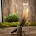 Load image into Gallery viewer, Drikkehorn Yggdrasil med Mønster, prydet med indviklede keltiske knudedesigns øverst, vises opretstående på en rustik træoverflade ved hjælp af et stativ. Grønt løv og mos i baggrunden skaber naturlige, jordnære omgivelser, der minder om et vikingemarked.