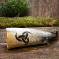 Load image into Gallery viewer, En Drikkehorn Triscele, prydet med et udskåret triquetra-symbol, ligger på en træoverflade dækket af mos. Grønt løv er let synligt i den slørede baggrund, hvilket skaber en rustik og naturlig indstilling, der minder om vikingemarkeder.