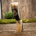 Load image into Gallery viewer, En håndlavet Drikkehorn Valknut med et slående mønster hviler oprejst på en træoverflade. Hornet har et sort og cremefarvet gradientdesign, prydet med et trekantet emblem. En naturholder støtter den på baggrund af grønne omgivelser og en rustik trævæg.