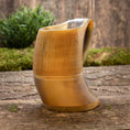 Load image into Gallery viewer, Et nærbillede af Vikingekrus Bark Horn (600-800ml) sat på en rustik træoverflade med grønt mos og planter i baggrunden. Ølkrusen viser en jævn gradient fra lys til mørk og har lodrette riller langs den øverste halvdel.