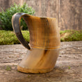 Load image into Gallery viewer, Et Vikingekrus Bark Horn (600-800ml) med træbund og håndtag hviler på en rustik træoverflade. Hornets krop er poleret med en naturlig gradient af lyse til mørkebrune nuancer, med udskårne lodrette linjer nær toppen. Grønne buske er slørede i baggrunden.