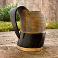 Load image into Gallery viewer, Et håndlavet Vikingekrus Bark Horn (600-800ml) med en poleret mørkebrun top og en lysebrun bund sidder på en rustik træoverflade med grønt i baggrunden. Denne ølkrus har et glat, buet håndtag og lodrette riller langs den øverste halvdel for tekstur.