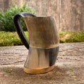 Load image into Gallery viewer, Et Vikingekrus Bark Horn (600-800ml) lavet af udskåret træ og poleret til en glat finish står på en rustik træoverflade. Hornet har lodrette riller langs kroppen, et sort håndtag og messingaccenter i bunden med grønt mos og træplader i baggrunden.