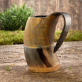 Load image into Gallery viewer, Et rustikt, håndlavet Vikingekrus Bark Horn (600-800ml) med lodrette kanter på den øverste halvdel og et glat, buet håndtag sidder på en træoverflade. I baggrunden er der grønt og en trævæg, der tilføjer en naturlig, jordnær følelse til scenen.