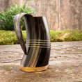 Load image into Gallery viewer, Et nærbillede af Vikingekrus Valknut Horn, et mørkt, vikinge-inspireret hornformet drikkekar med udskåret håndtag og vandrette dekorative linjer. Basen ser ud til at være lavet af træ. Baggrunden har grønt løv og en rustik træoverflade, der tilføjer den historiske charme af dette unikke drikkekar.