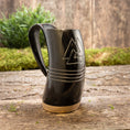 Load image into Gallery viewer, Et Vikingekrus Valknut Horn (600-800ml), sort og minder om vikingetraditioner, sidder på en træbase på en rustik træoverflade. Hornet er indgraveret med et trekantet design og tre vandrette linjer. I baggrunden er der grønt og en trævæg, der forstærker den naturlige og rustikke stemning.