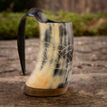 Load image into Gallery viewer, En Mini Vikingekrus Horn Wave (100 ml) med indviklede keltiske knudeudskæringer og et glat håndtag sidder på en rustik træoverflade. Hornkrusen fremviser en blanding af sorte, hvide og beige nuancer, og dens flade base gør, at den kan stå oprejst.
