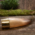Load image into Gallery viewer, En Drikkehorn Elegance med indviklet metalarbejde i den brede ende er placeret på en rustik træoverflade. Bagved formidler et lag grønt mos og sløret grønt en naturlig og historisk atmosfære, der minder om vikingemarkeder.