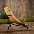 Load image into Gallery viewer, Drikkehorn Elegance, prydet med indviklede endestykker af guld, hviler yndefuldt i et slankt sort smedejernsstativ. Set mod en rustik trækulisse og omgivet af grønne områder fremhæves hornets håndværk og historiske charme smukt, hvilket gør det til et ideelt stykke for vikingemarkeder.