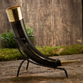 Load image into Gallery viewer, Drikkehorn Elegance, et dekorativt drikkehorn, holdt i et udsmykket metalstativ prydet med indviklede guldudsmykninger i begge ender, er sat mod en rustik træbaggrund med grønt mos og løv. Denne scene fremkalder vikingemarkedernes vintage charme og en middelalderlig stemning.
