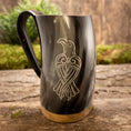 Load image into Gallery viewer, Et poleret Vikingekrus Valravnen Horn (600-800 ml) har et indgraveret design af en stiliseret fugl på siden. Tankarden viser et sort håndtag og en metallisk base, der hviler elegant på en rustik træoverflade med grønt mos i baggrunden.