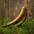 Load image into Gallery viewer, En poleret, buet Drikkehorn Devine med udsmykkede metalbånd i begge ender hviler på en plet af grønt græs. Hornet, der minder om dem, man fandt på vikingemarkeder, er støttet op med en sort rem, og en rustik trækulisse ses i baggrunden.
