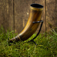 Load image into Gallery viewer, Drikkehorn Devine, et poleret og elegant buet drinkhorn prydet med dekorative bånd på kanten og bunden, er fremvist på et elegant sort metalstativ. Set mod grønt græs med et rustikt træhegn i baggrunden, fremkalder scenen levende billeder af travle vikingemarkeder.