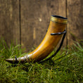 Load image into Gallery viewer, En Drikkehorn Devine med elegante metalaccenter hviler på en græsklædt overflade. Hornet er placeret oprejst og understøttet af et sort metalstativ, sat mod den rustikke kulisse af en trævæg, der minder om scener fra vikingemarkeder.