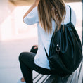 Load image into Gallery viewer, En person med langt hår og en hvid skjorte sidder på en bænk og vender væk. De bærer sorte jeans og bærer en sort rygsæk med snoretræk med Gede Kaffe Drikkehorn Sort påklipnings BPA-fri vandflaske. Baggrunden er sløret, hvilket antyder en udendørs bymiljø.