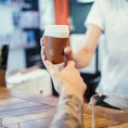 Load image into Gallery viewer, En tatoveret arm rækker en hånd ud for at tage imod en ged Kaffe Drikkehorn Brun fra en barista på tværs af cafedisken, der minder om en moderne viking, der handler med sit drikkehorn. Baggrunden er sløret og fokuserer scenen på kaffeoverdragelsen.