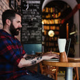 Load image into Gallery viewer, En skægget mand med tatoveringer, der ligner en moderne viking, er iført en rød og blå plaid skjorte, mens han taster på en bærbar computer ved et rundt træbord i en hyggelig café med bogreoler og tavlevægge. En ged Kaffe Drikkehorn Brun isoleret kop lavet af BPA-fri plastik sidder tæt på hans bærbare computer.