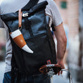 Load image into Gallery viewer, En person iført en grå t-shirt bærer en stor sort roll-top rygsæk med en hvid og solbrun ged Kaffe Drikkehorn Brun påsat, mens han skubber en cykel med en brun Brooks-sadel i læder. Scenen fremkalder stemningen fra en moderne viking på en bygade.