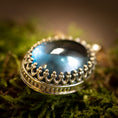 Load image into Gallery viewer, Et nærbillede af et udsmykket sølvvedhæng med en stor, blank London Blue Topaz i midten, hvilende på en bund af grønt mos. Vedhæng - Eryn Design - Tranquill 14kt Guld vedhænget er designet af Eryn Design og har indviklede detaljer, der ligner små kroner rundt om ædelstenen, hvilket skaber et kongeligt og elegant udseende.
