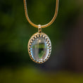 Load image into Gallery viewer, Et nærbillede af et elegant vedhæng hængende fra en guldkæde. Vedhæng - Eryn Design - Tranquill 14kt Guld vedhæng har en ovalformet London Blue Topas indkapslet i en udsmykket 14kt guldindfatning med indviklede detaljer. Baggrunden er sløret grønt, hvilket tilføjer en naturlig, frodig kontekst.
