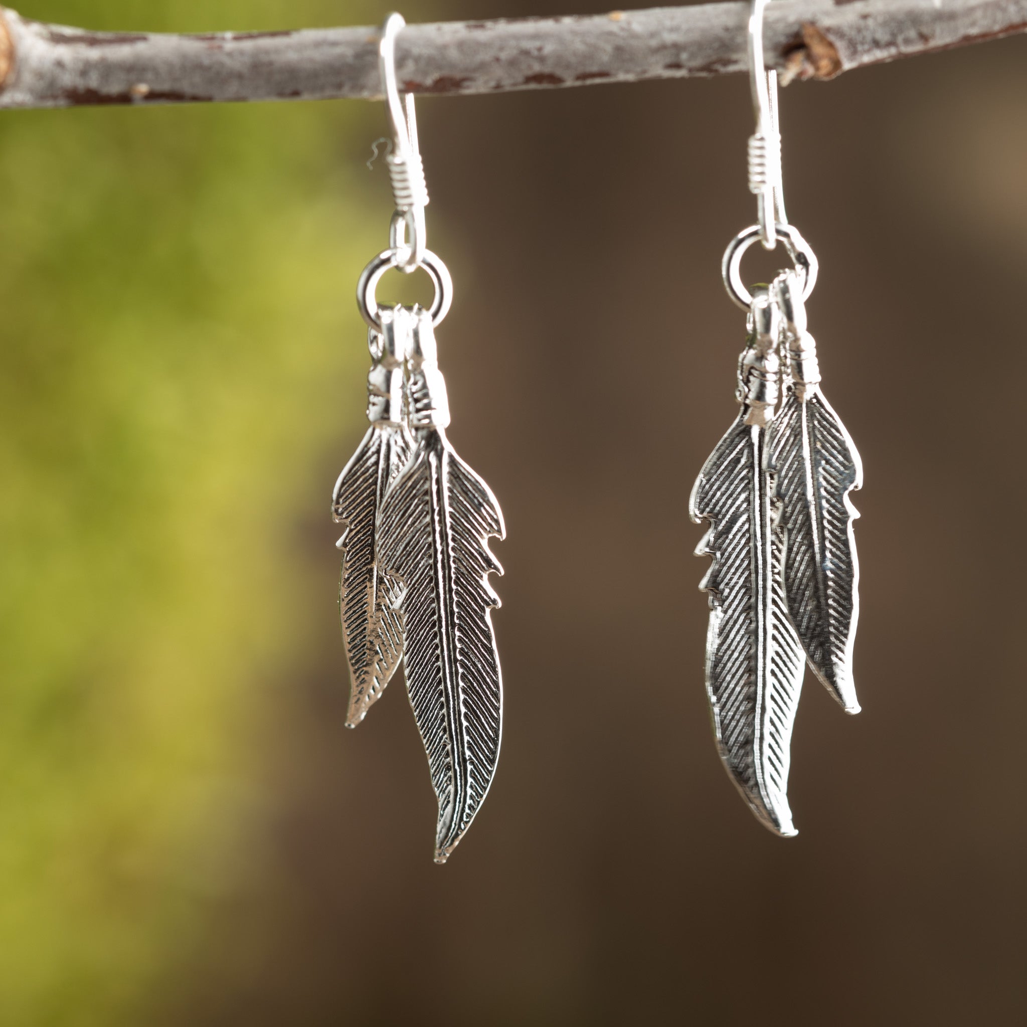 Nærbillede af et par Hængeøreringe Feather 925s Sølv, sart hængende fra en tynd trægren. Disse fantastiske øreringe kan prale af indviklede detaljer, der ligner ægte fjer, og fanger naturlige motiver og er sat mod en sløret, jordagtig baggrund med grønne nuancer.