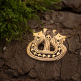 Load image into Gallery viewer, Et Broche Vikingeskib Bronze med indviklede designs af to stiliserede hestehoveder og et centralt kranium hviler på en revnet, jordagtig overflade. Grønt mos er synligt i venstre side af billedet. Brochen fremkalder udseendet af en middelalderlig eller gammel vikingetiden-artefakt.
