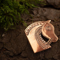 Load image into Gallery viewer, En bronze, indviklet designet Broche Hest fra Sösdala Bronze, der forestiller et hestehoved, ligger på revnet jord ved siden af en lille plet grønt mos. Detaljerne på brochen inkluderer udsmykkede mønstre og et stiliseret øje, der viser håndværk inspireret af det nomadiske hestefolk.
