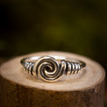 Load image into Gallery viewer, Sølvring Spiral 925s Sølv er elegant placeret på en træoverflade. Denne sølvring kan prale af et slående spiraldesign i midten, med indviklet wirework, der fremhæver spiralen, der afspejler vikingernes håndværk. Baggrunden er blødt sløret med jordnære og grønne nuancer.
