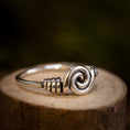 Load image into Gallery viewer, Et nærbillede af Sølvring Spiral 925s Sølv-ringen med et indviklet oprullet tråddesign i midten, der minder om Vikingerne håndværk. Ringen er placeret på en træoverflade med en mørk, sløret baggrund, der fremhæver ringens håndværk og skinnende, reflekterende egenskaber.
