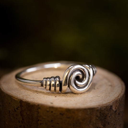 Et nærbillede af Sølvring Spiral 925s Sølv-ringen med et indviklet oprullet tråddesign i midten, der minder om Vikingerne håndværk. Ringen er placeret på en træoverflade med en mørk, sløret baggrund, der fremhæver ringens håndværk og skinnende, reflekterende egenskaber.