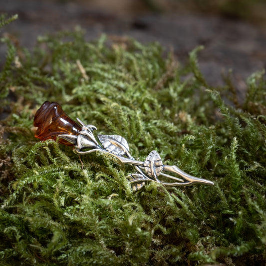 Rose Brooch with Amber 925s Silver, a silver brooch shaped like a stem with leaves, holds authentic amber like a flower bud and rests on vivid green moss. Its detailed craftsmanship mimics a natural rose.