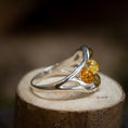 Load image into Gallery viewer, Et nærbillede af Sølvring Amber Drops 925s Sølv, en smukt udformet sølvring med et kunstnerisk, flydende design med tre runde ravsten i gule, orange og grønne nuancer. Ringen vises oven på en lille træstub, der fremhæver dens detaljerede håndværk mod en blødt sløret baggrund.
